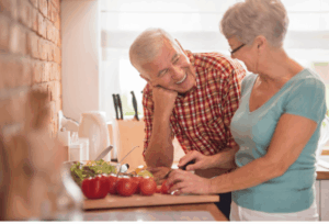 Couple partageant un moment convivial dans une cuisine, un homme riant avec sa femme qui prépare des tomates, illustrant le plaisir des échanges et le confort d’écoute au quotidien.